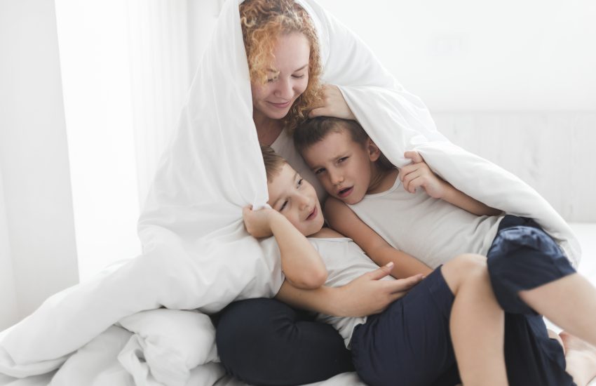 Mama bawi się z dwójką dzieci pod kołdrą w przytulnym pokoju z bezpiecznym piętrowym łóżkiem dla dziecka w tle.