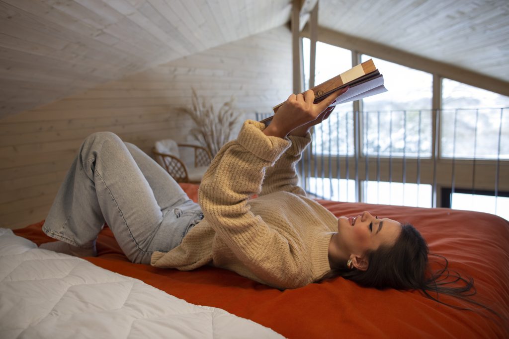 Młoda kobieta czyta książkę, leżąc wygodnie na łóżku na antresoli w przytulnym, drewnianym wnętrzu