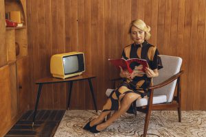Kobieta w stylizacji retro czyta magazyn, siedząc w fotelu w salonie urządzonym w stylu mid-century modern, gdzie widoczne są charakterystyczne drewniane meble lata 60., w tym trójkątny stolik pod żółty telewizor i regał.