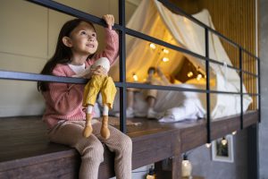 Dziewczynka z lalką siedzi przy balustradzie, a w tle widać oświetlone łóżko na antresoli z zadaszoną konstrukcją z tkaniny
