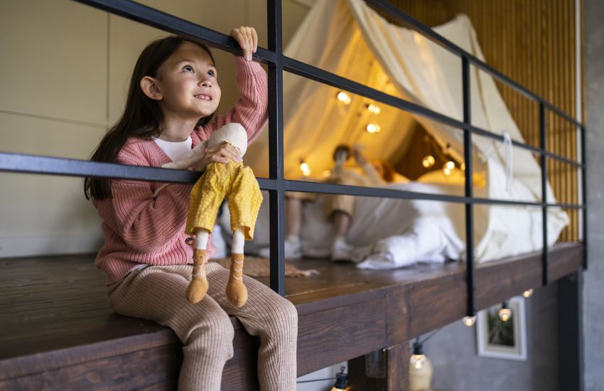 Dziewczynka z lalką siedzi przy balustradzie, a w tle widać oświetlone łóżko na antresoli z zadaszoną konstrukcją z tkaniny