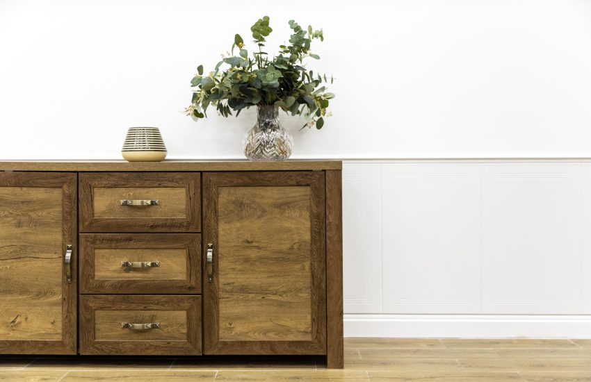 Rustykalna komoda z ciemnego drewna z trzema szufladami, szafkami i mosiężnymi uchwytami, udekorowana wazonem z eukaliptusem na tle białej boazerii.