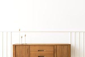 Drewniana komoda w stylu mid-century modern z charakterystycznymi ryflowanymi drzwiczkami i gładkimi szufladami, na której stoją trzy złote świeczniki.