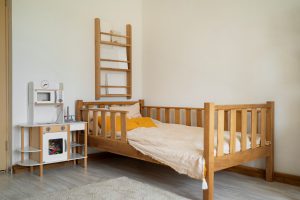 Łóżka dziecięce 140x70 z pionowymi szczebelkami pełniącymi funkcję barierki ochronnej. Łóżko jest zasłane jasną pościelą z żółtymi poduszkami dekoracyjnymi