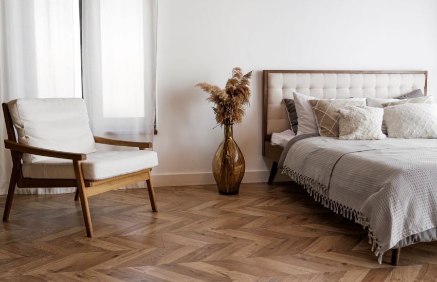 Jasna i przytulna sypialnia po zakończeniu prac, które obejmowały kompleksowy remont sypialni. Na pierwszym planie widać dębowy parkiet ułożony w jodełkę oraz biały fotel, a w tle duże łóżko z tapicerowanym zagłówkiem i szarym pledem.