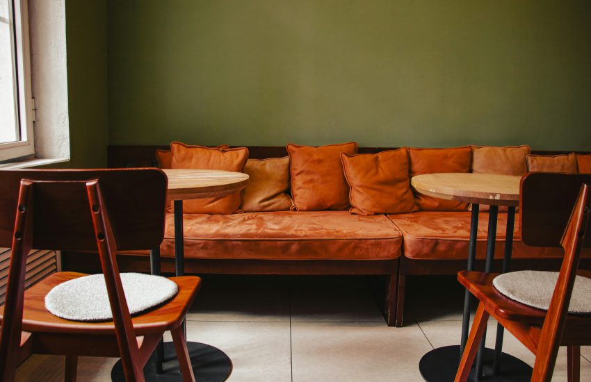 Na zdjęciu widać przytulne wnętrze kawiarni z długą sofą w ciepłym, rdzawym odcieniu ustawioną przy zielonej ścianie