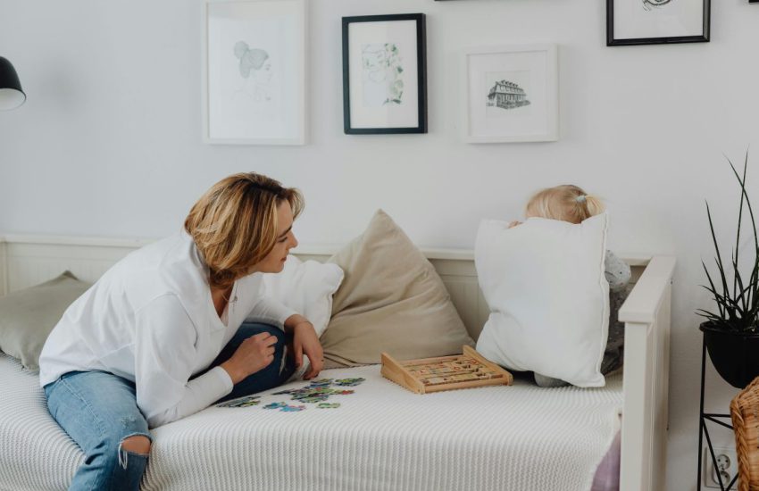 Kobieta i dziecko bawią się na białym tapczanie z funkcją spania, ozdobionym beżowymi poduszkami, w jasnym pokoju z obrazkami na ścianie.